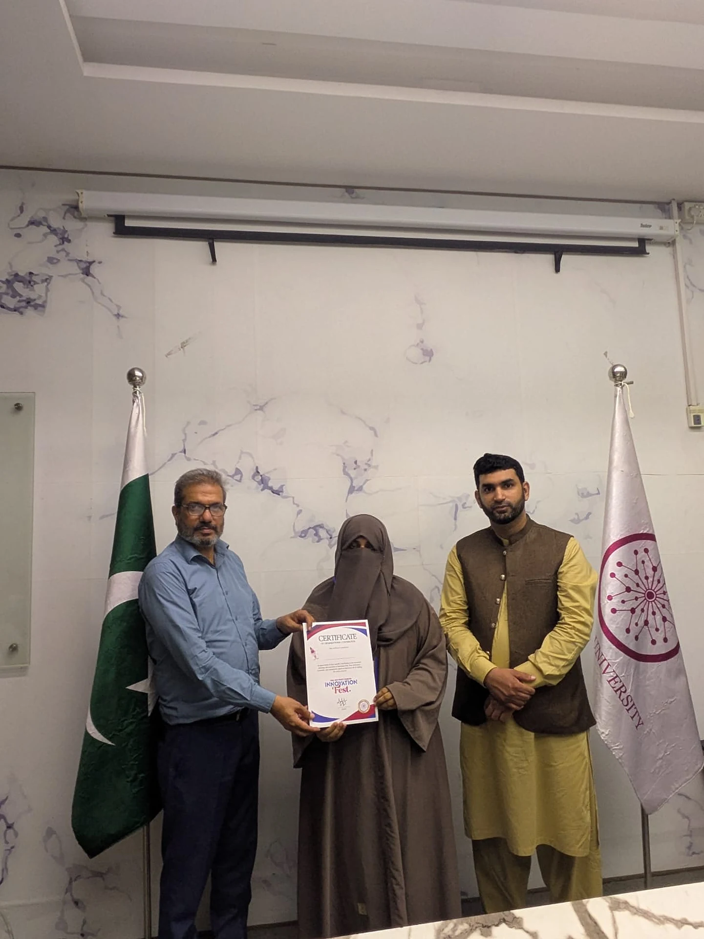 A certificate presentation taking place between three people beside two standing flags.