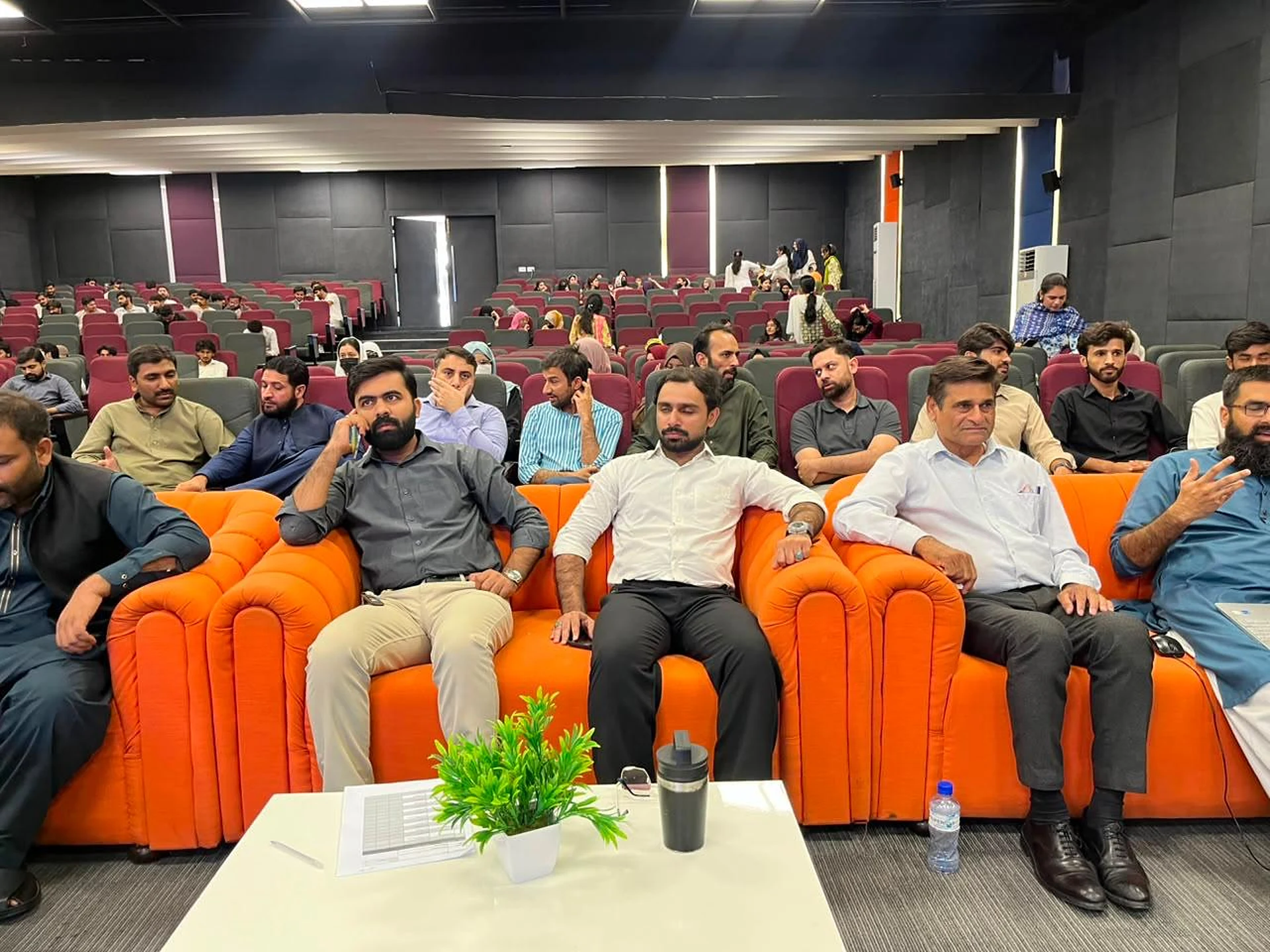 Participants seated on bright orange sofas at the front of an auditorium.