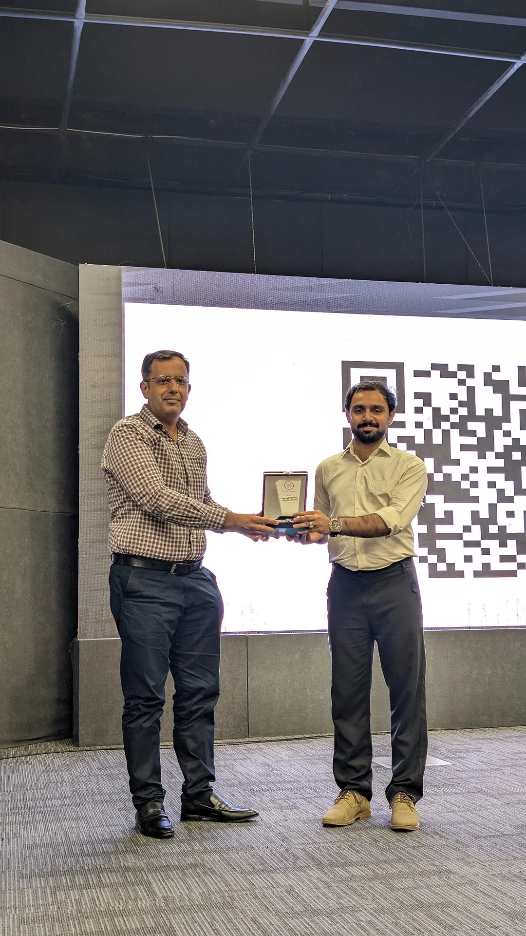 Two people on stage holding an award plaque in front of a large screen.